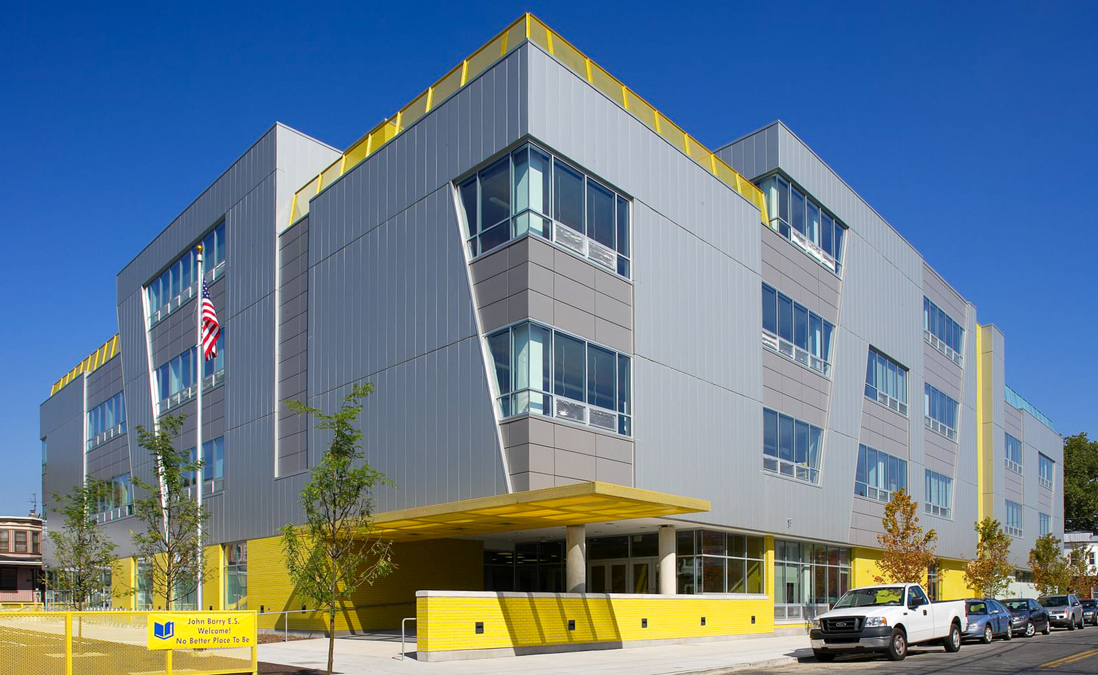 Exterior view of Commodore John Barry Elementary School in Philadelphia, showing the modern gray and yellow building with large windows, a bright yellow entrance, and an American flag displayed in front.